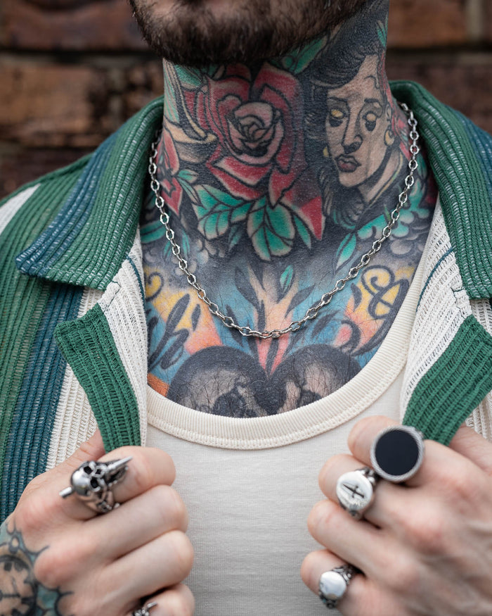 A close-up of a tattooed neck and chest with colorful rose and woman's face tattoos. The person wears silver rings, a white shirt, green striped jacket, and "The Cathedral" 6mm Spiked Chain necklace.