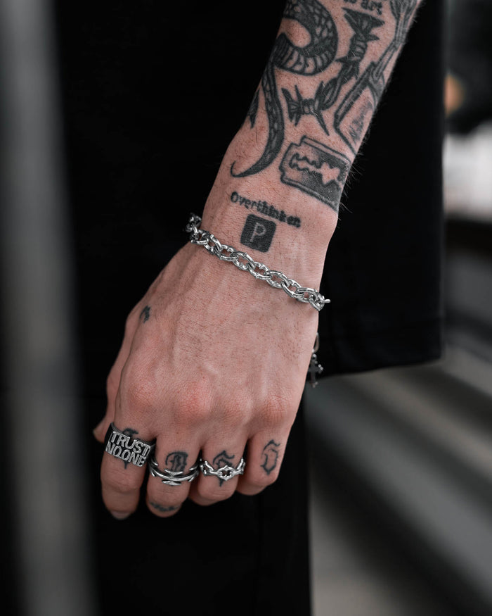 A tattooed hand adorned with silver rings and "The Cathedral" 9mm Spiked Bracelet, a non-tarnish waterproof medieval chain. Tattoos read Overthate, a boxed P, and TRUST NO ONE across the fingers. The individual wears black attire.