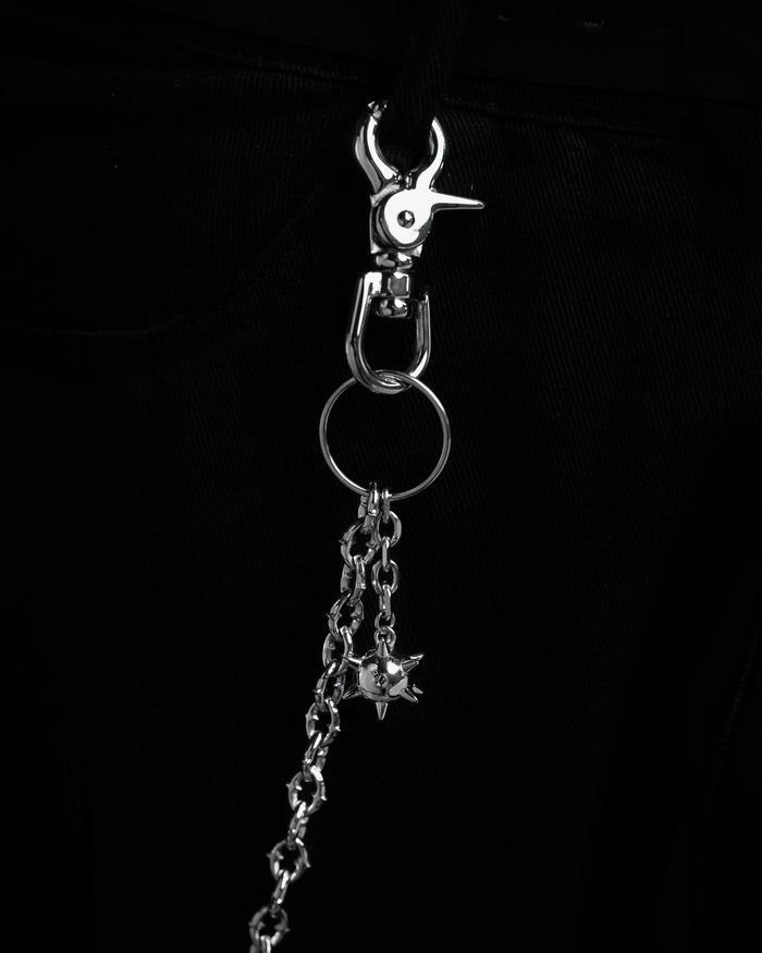 A close-up of "The Cathedral" 9mm Spiked Wallet Chain with a ball charm, metal clip, and key ring, hanging from black fabric—possibly pants. The dark, monochromatic look adds to its edgy streetwear style.