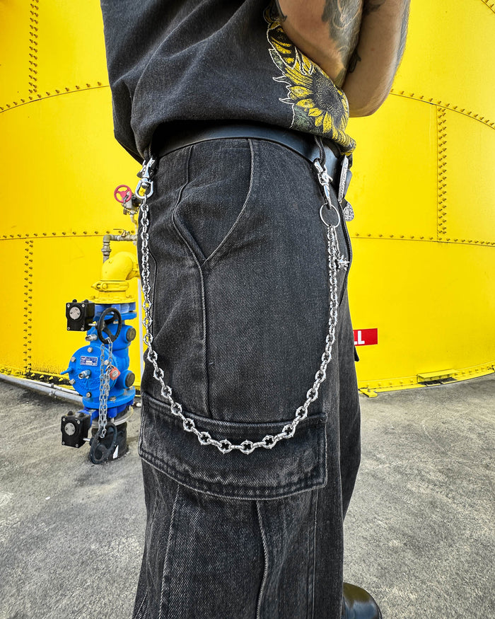 A person in black cargo pants with "The Cathedral" 9mm Spiked Wallet Chain clipped to their belt loop stands before a large yellow industrial tank, with blue and yellow valves visible in the background.