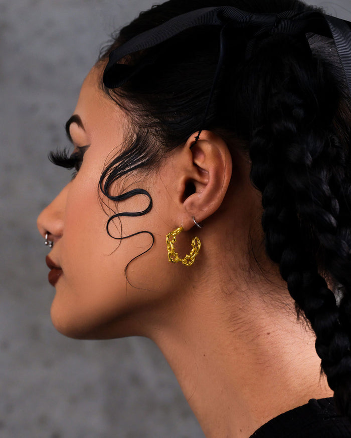 A person with braided hair, a black ribbon, and a face-framing curl wears "The Cathedral" Hoop Earrings (Gold) and a nose ring, shown in profile against a gray background.
