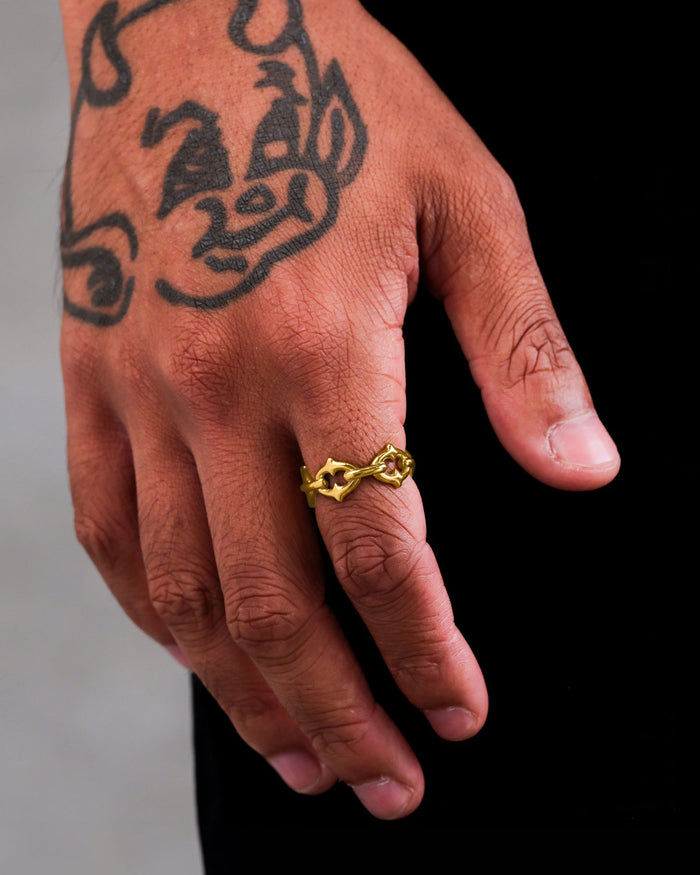 A hand with a cartoon-style devil tattoo wears Statement Collective’s "The Cathedral" Spiked Ring (Gold)—a chunky, twisted 18k gold piece—on the ring finger. The plain dark background highlights its bold elegance.