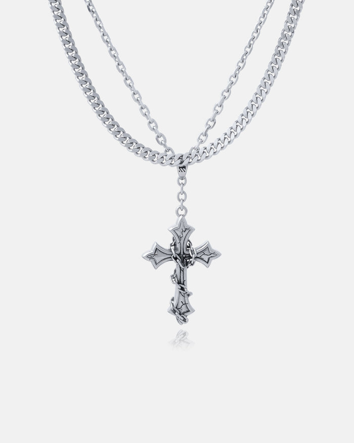 The Ruins" 6mm Set features a layered stainless steel necklace with a gothic cross pendant on the shorter chain and a Cuban link chain, all showcased against a plain white background.