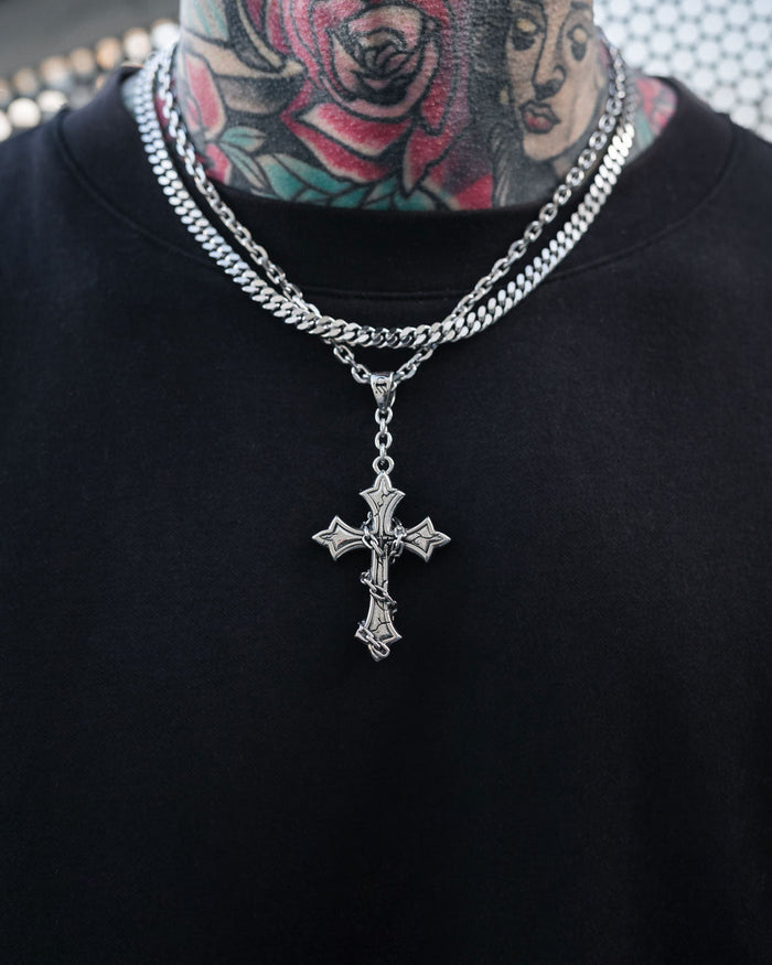 A person wearing a black shirt and Statement Collective's "The Ruins" 6mm Set, featuring layered stainless steel jewelry with a gothic cross pendant on a cuban link chain, showing off colorful floral and face tattoos on their neck.
