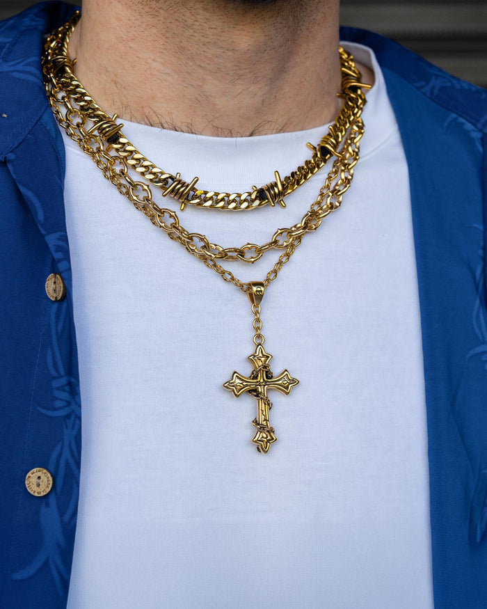A person wearing layered gold chains from Statement Collective, including the Spite Set Gold (Custom Pendant Choice) featuring an 18k gold chain with a cross pendant and a custom pendant necklace, over a white T-shirt and a blue button-up shirt from the Spite Stack collection.