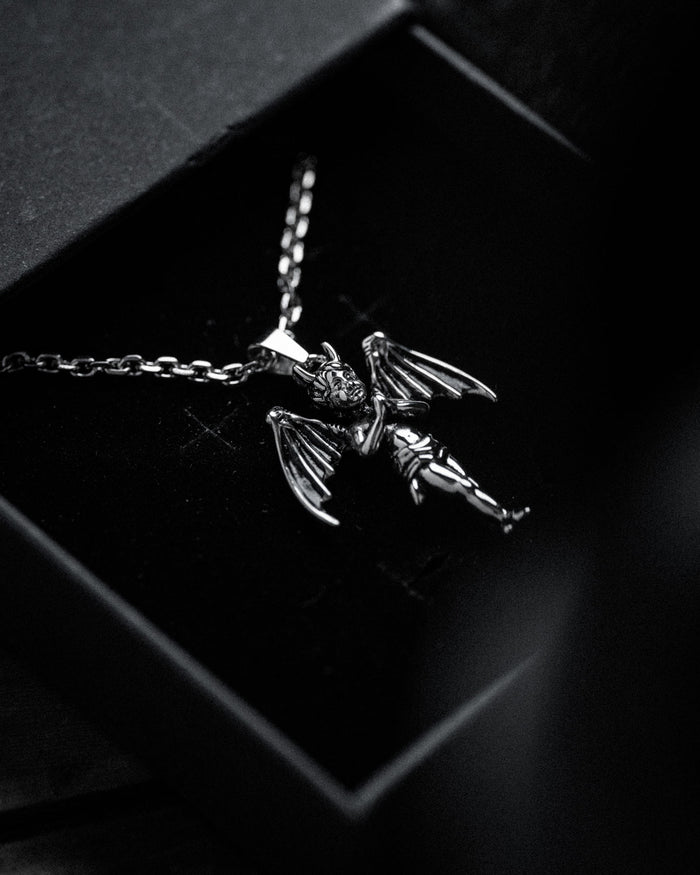 A black and white close-up of "The Wicked One" Pendant, a gothic necklace with a winged, horned cherub pendant, shown inside a black jewelry box to highlight its intricate details.